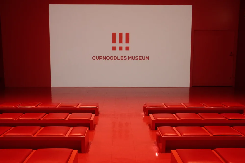 A red auditorium with rows of benches and a screen displaying the Cup Noodles Museum logo